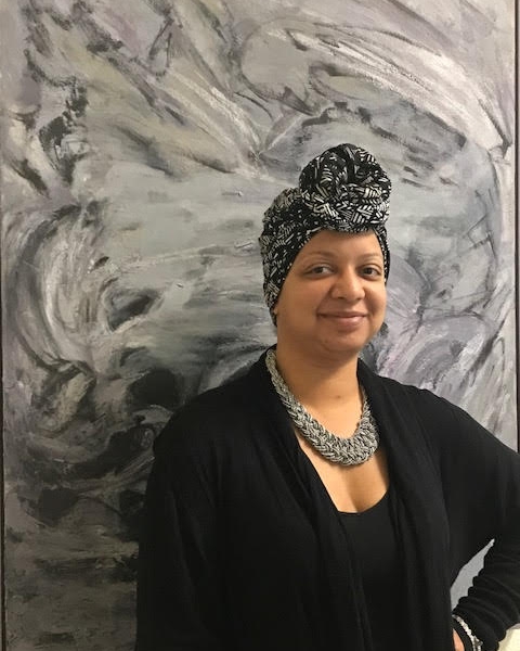An African American woman wearing a grey head wrap and black cardigan poses in front of a grey marble like background for her portrait.