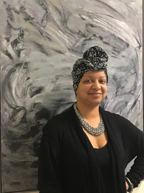 An African American woman wearing a grey head wrap and black cardigan poses in front of a grey marble like background for her portrait.
