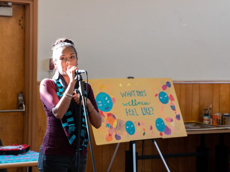 A young indigenous woman with her hair in a bun speaks in a microphone at a well-lit room. A sign behind her says "What Does Wellness Feel Like?"