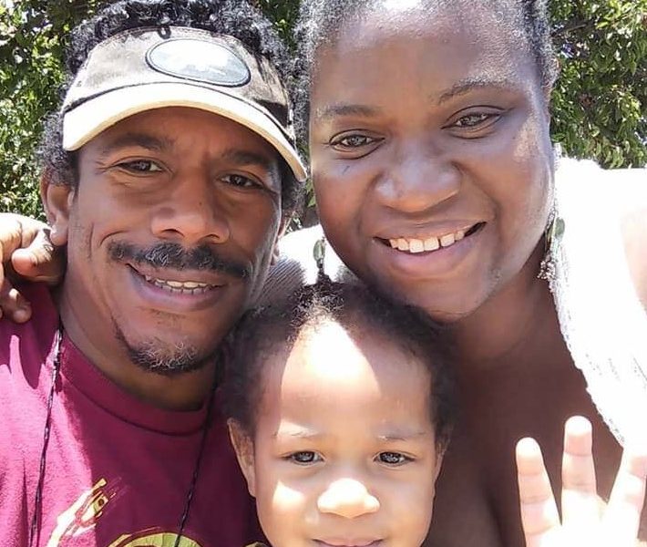 An African American family (man, woman, and toddler) smile at the camera in a selfie photo.