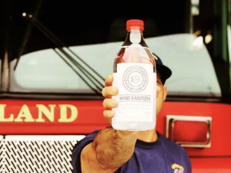 A firefighter is holding a glass container of hand sanitizer. In the back is a bright red firetruck that says "Oakland Fire Dept."