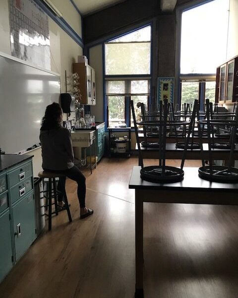 A dark image of a high school classroom that is empty except the teacher.