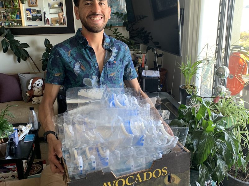A smiling man carries a cardboard box filled with plastic face shields for Kaiser nurses. The cardboard box says "avocados" on it.