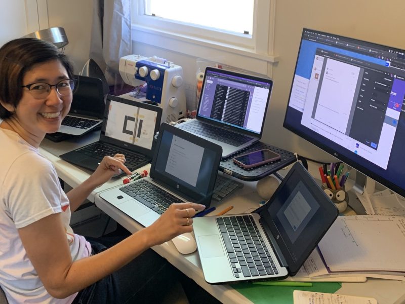 A smiling East Asian woman with a bob haircut and glasses is sitting at a large desk with several Chromebook laptops in front of her, plus a big monitor.