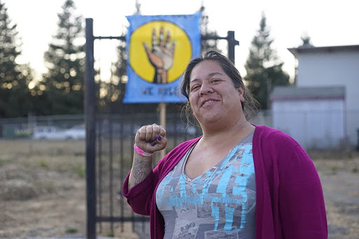 A woman smiles and holds up one fist in front of a housing encampment in East Oakland.