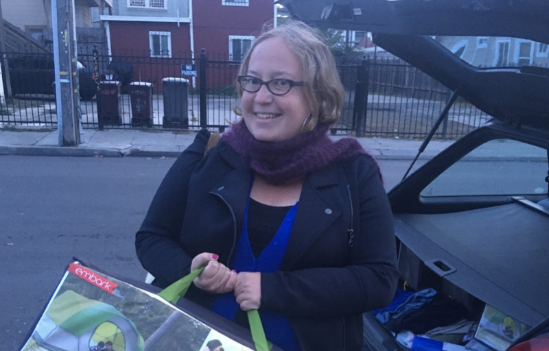 a woman with blonde hair and glasses smiles at the camera in front of a car.