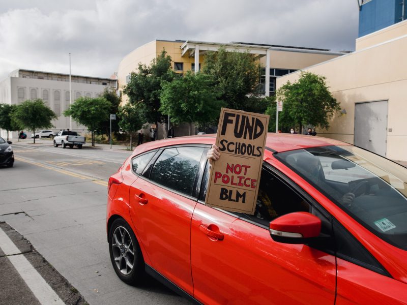 A red car has the window open. A hand is holding a homemade cardboard sign that says "Fund Schools Not Police BLM"