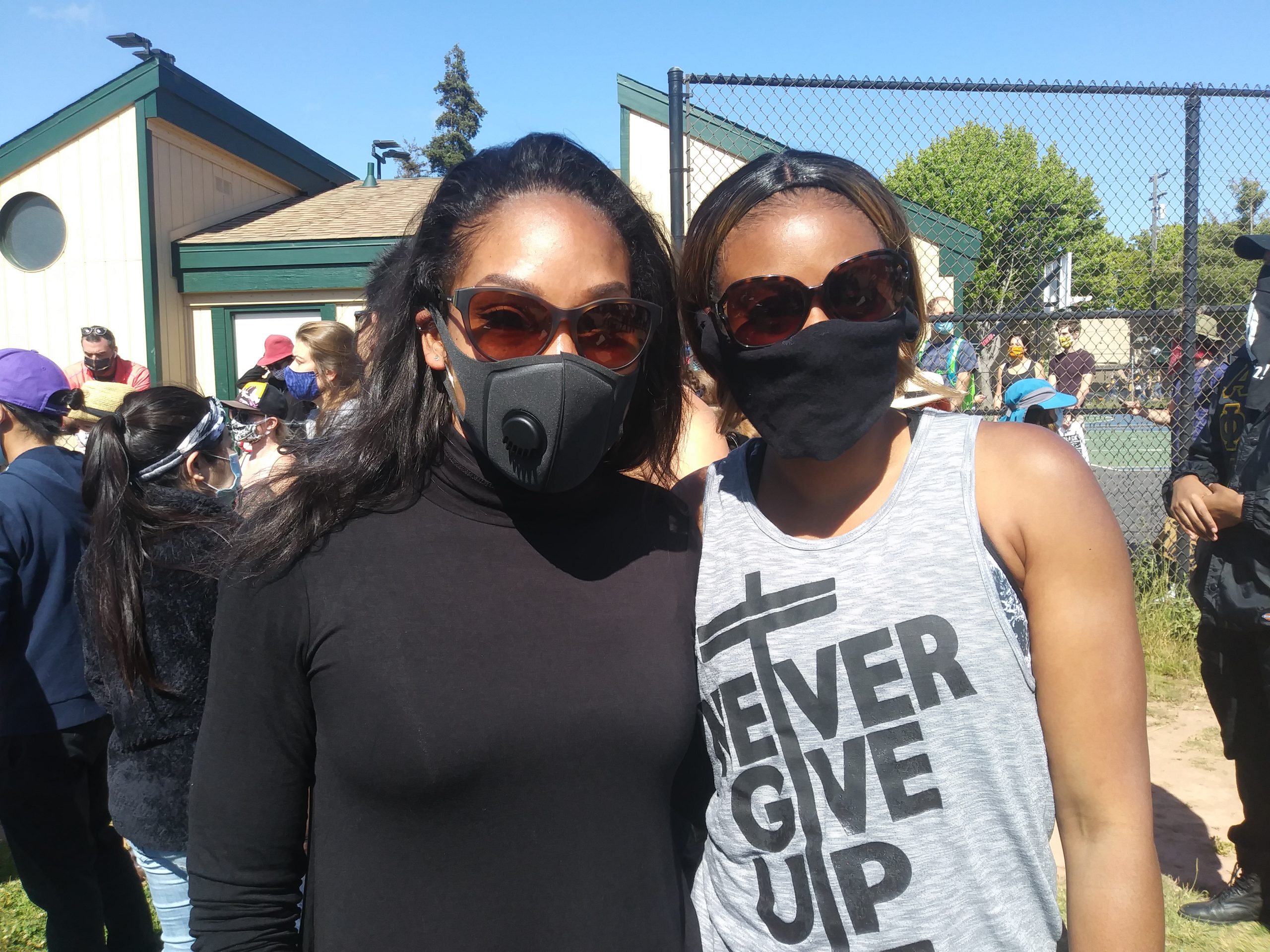 Two Black women wearing black face masks stand next to each other and pose for a photo. The sun is bright.