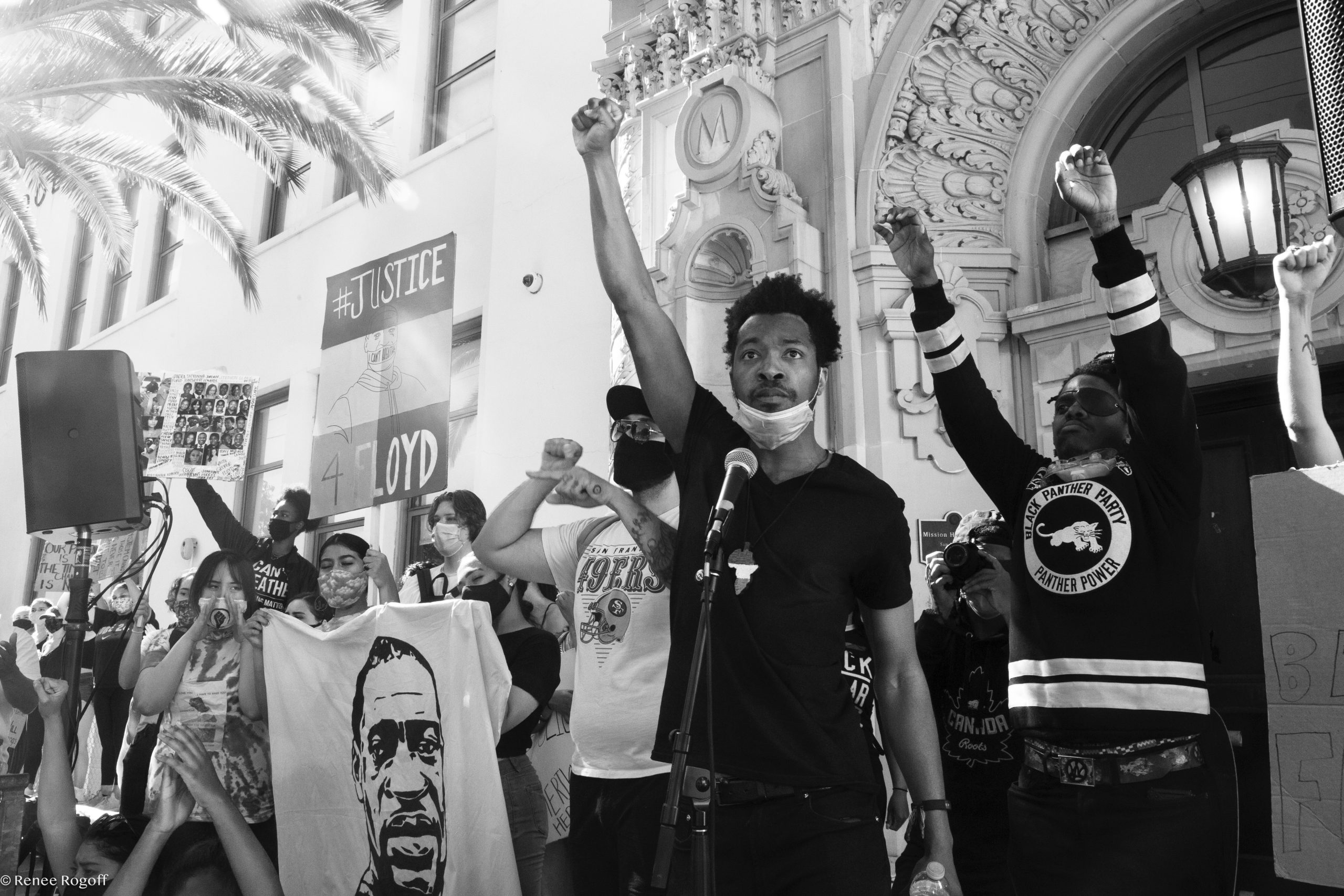 A Black man raises his fist in the air in front of an ornamental building. The photo is in black and white.
