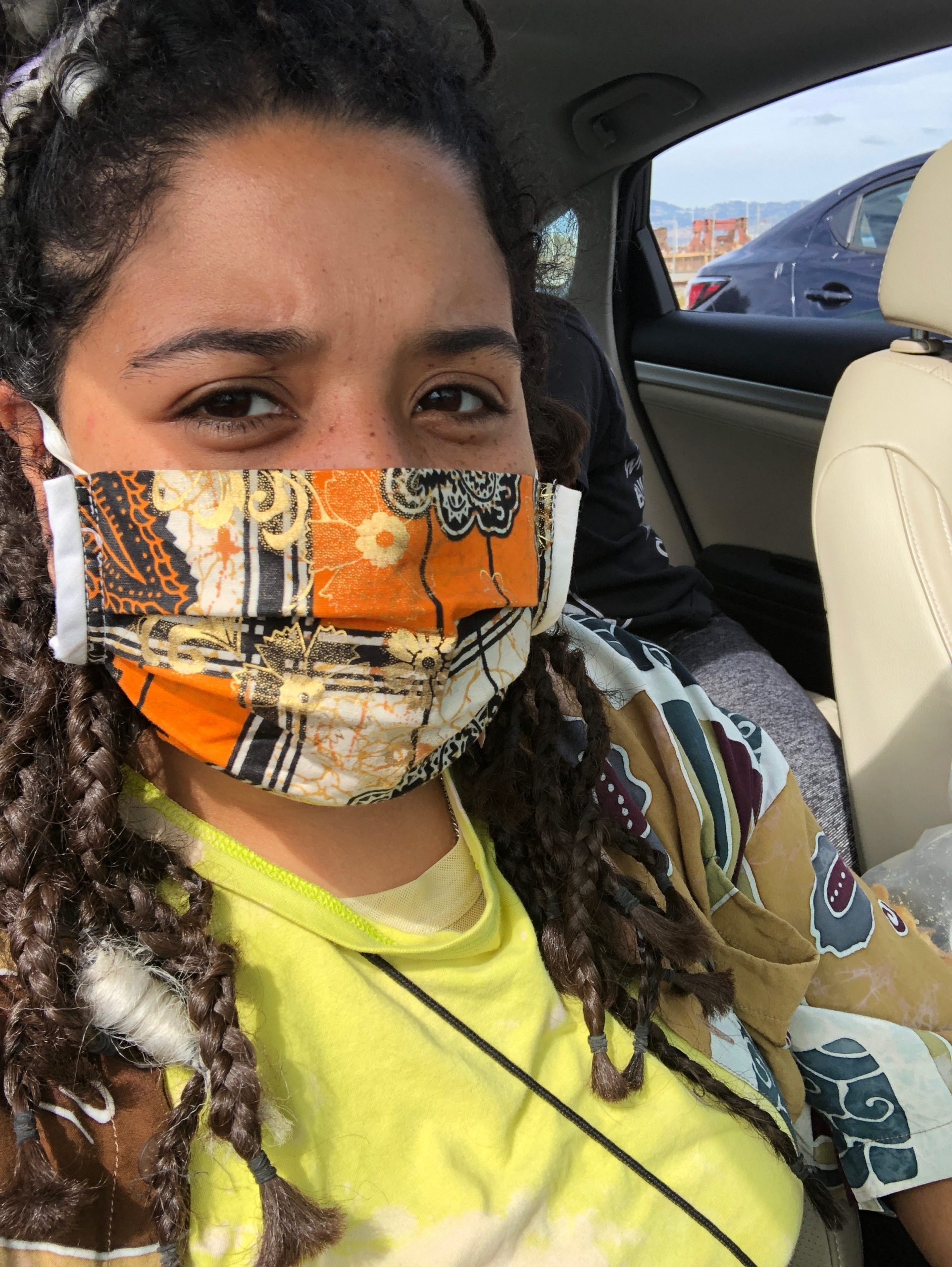 A biracial woman with braids sits in a car during a protest and is wearing a colorful orange and black mask.
