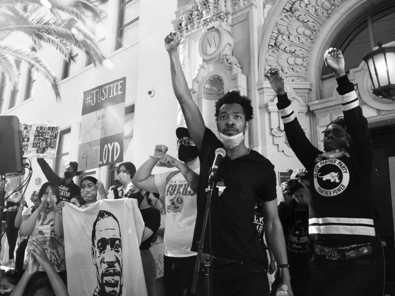 A Black man raises his fist in the air in front of an ornamental building. The photo is in black and white.