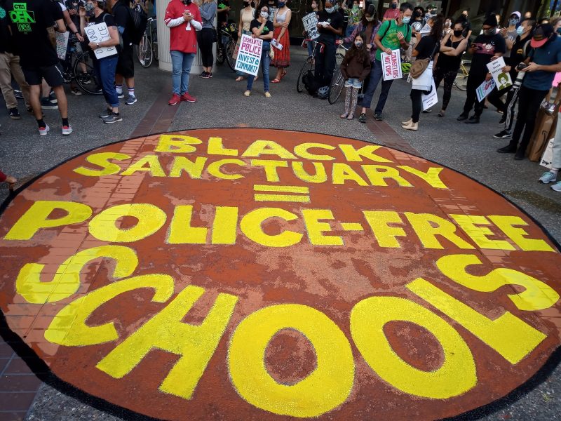 A sign painted on the ground reads "Black sanctuary = police-free schools" with people in masks behind the sign.