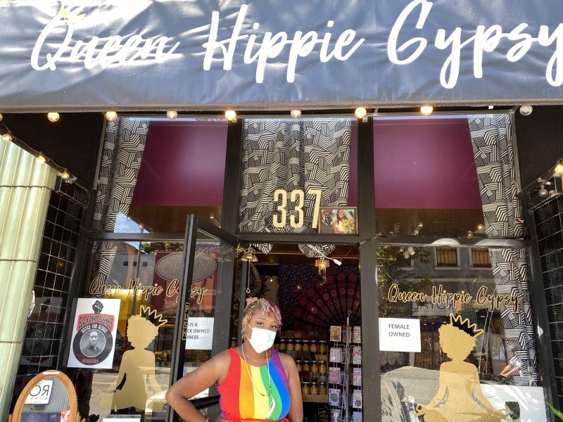 An African American woman wearing a rainbow colored dress stands in front of a storefront in Oakland.
