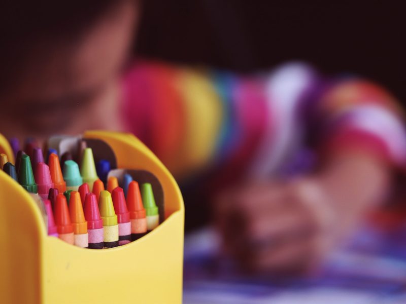 A closeup of crayons and a child in the background coloring.