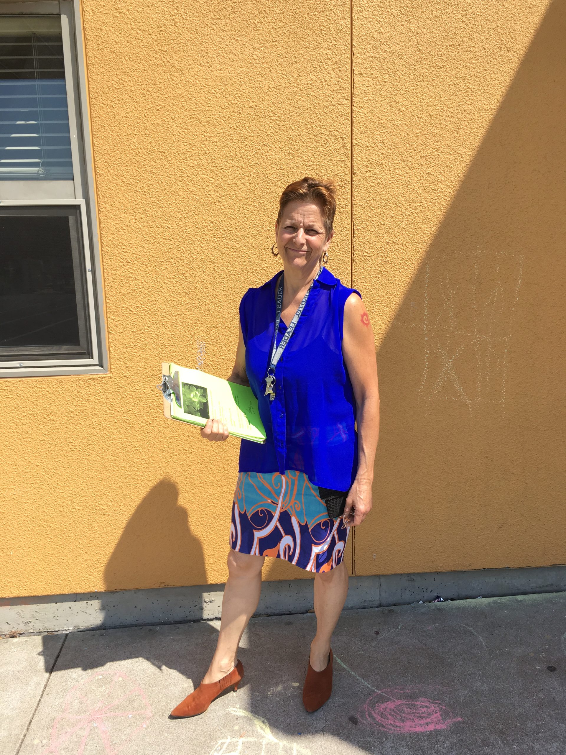 A woman with short hair and bright blue blouse stands in front of a school.