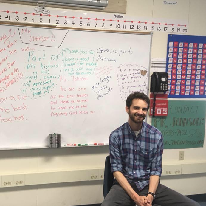A teacher sits in a chair in front of a whiteboard.