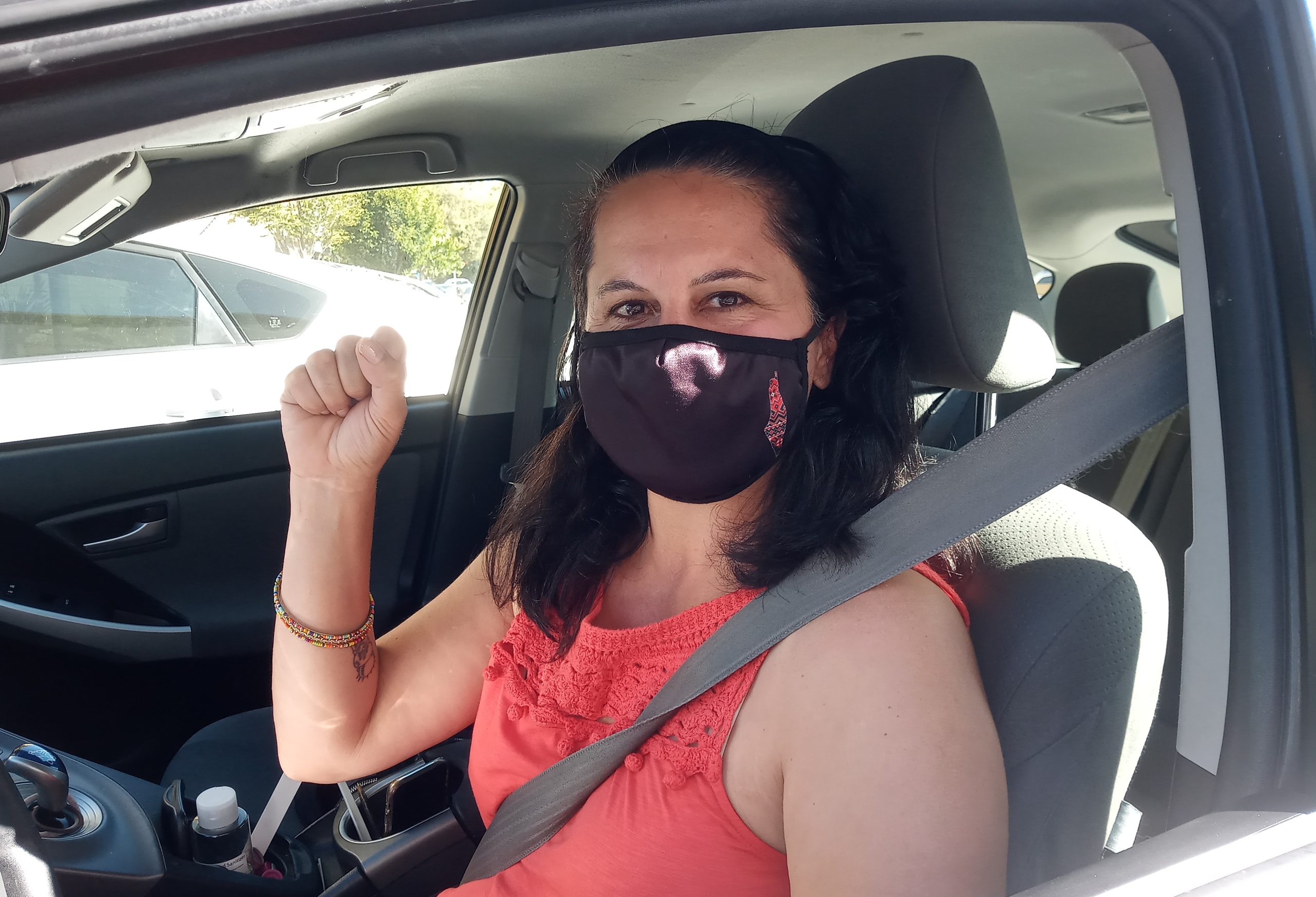 Noura Khouri. A woran wearing a facemask and her fist up sitting inside her car.