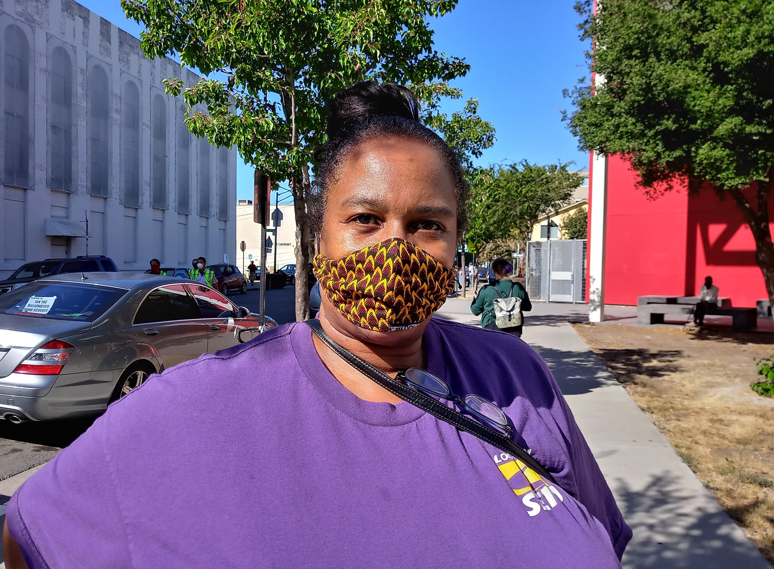 An African American woman wearing a facemark and purple t-shirt looks at camera.