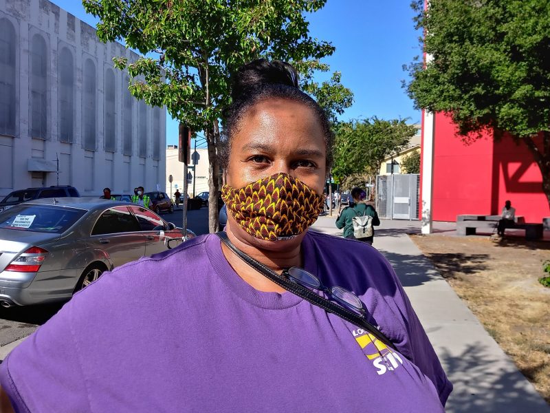 An African American woman wearing a facemark and purple t-shirt looks at camera.