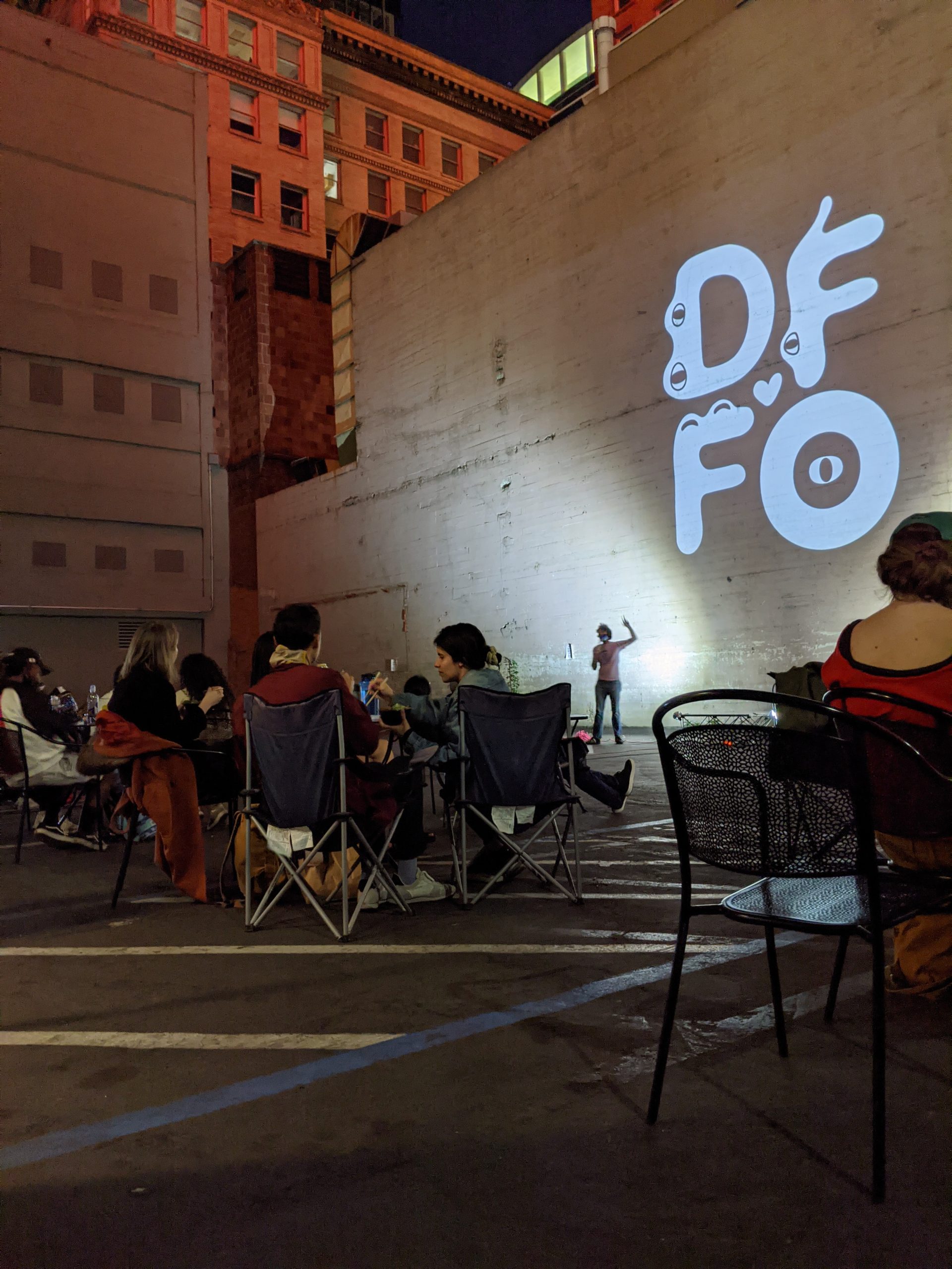 An image of a man in a white t-shirt against a wall in the evening while patrons sit in small clusters in a parking lot.