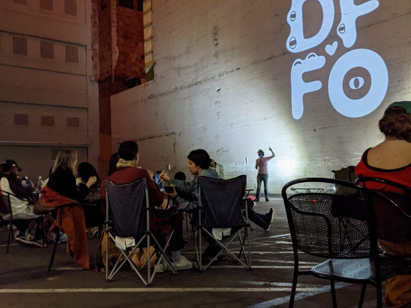 An image of a man in a white t-shirt against a wall in the evening while patrons sit in small clusters in a parking lot.