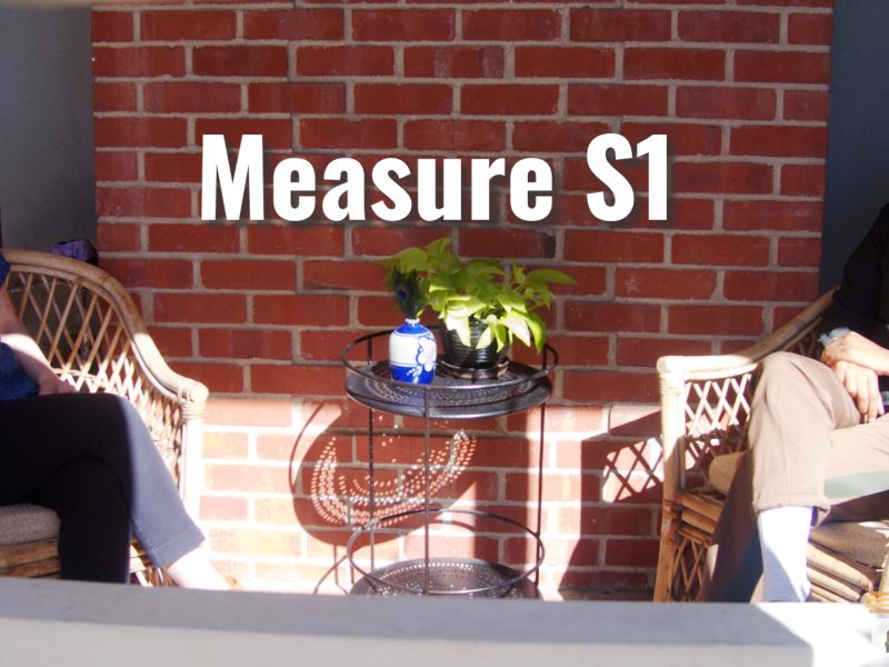 A woman wearing a mask sits on a sunlit porch with a woman wearing a mask.