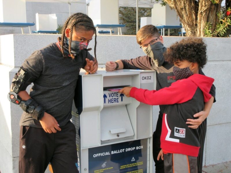 A mom and two children stand by a ballot box dropping off their ballot.