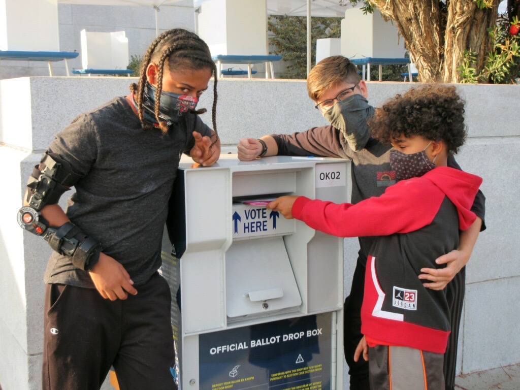 A mom and two children stand by a ballot box dropping off their ballot.