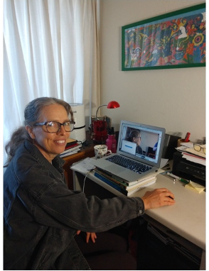 A woman wearing glasses and grey hair in a pony tail smiles for camera while sitting at desk.