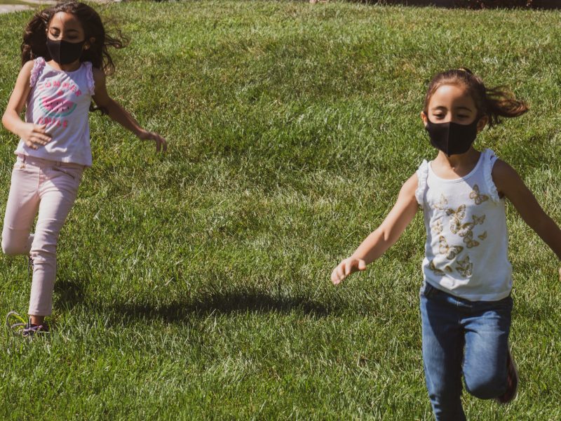 Two girls running on grass wearing black face masks.