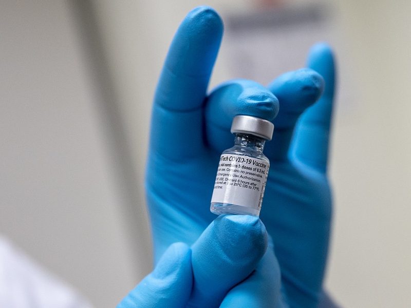 An image of a hand in blue gloves holding a vial of the COVID-19 vaccine.