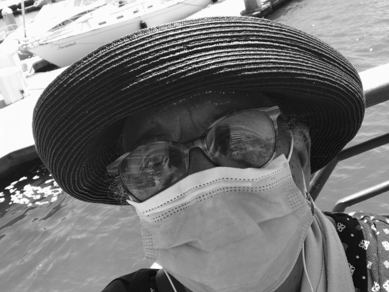 A black and white image of a woman wearing a hat, glasses, and a face mask standing outside by boat docks