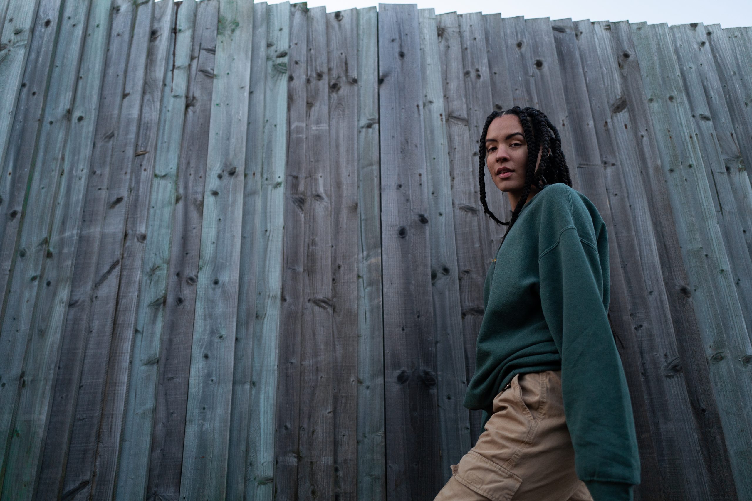 An image of an African American woman walking in front of a fence, with long braids and a green sweatshirt. She is smiling.
