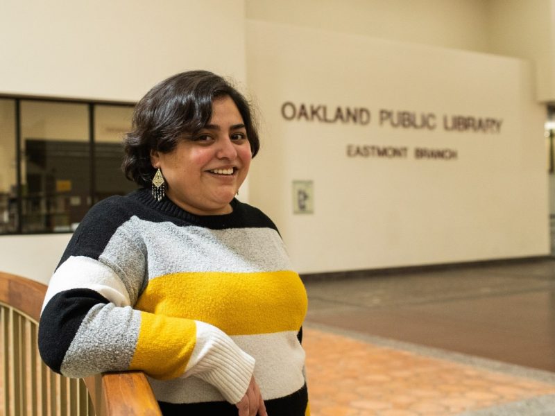 A Guatemalan American young woman with short, wavy hair stands in front of a sign that says Eastmont Library