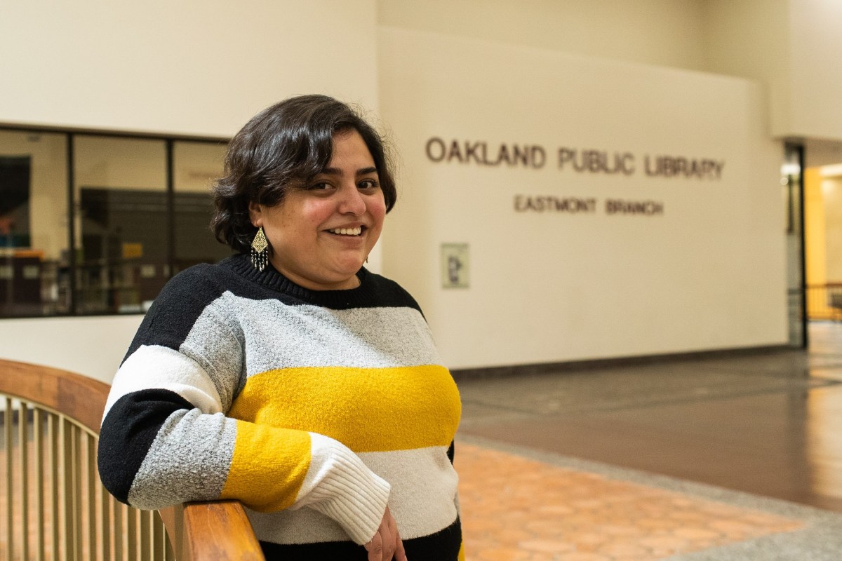 A Guatemalan American young woman with short, wavy hair stands in front of a sign that says Eastmont Library