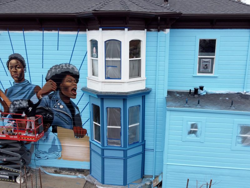 An image of a Victorian home painted with baby blue and images of powerful Black women from the Black Panther Party painted on the outside of the house in West Oakland.