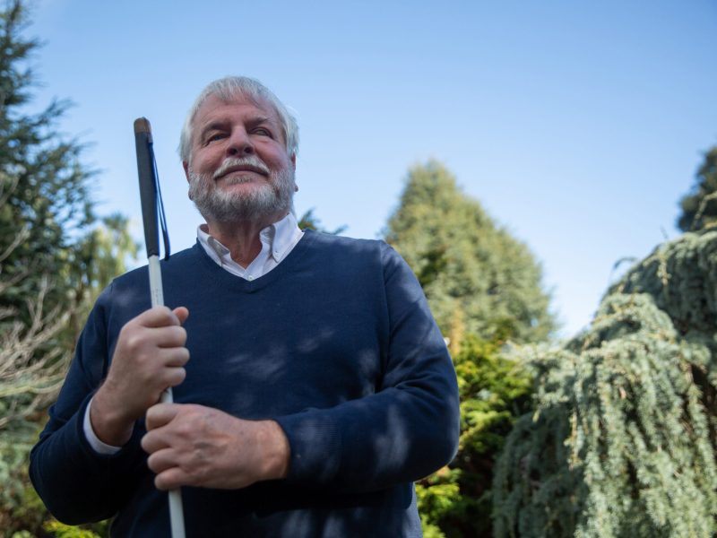 An image of a man who is blind with short grey hair, and green trees in the background.