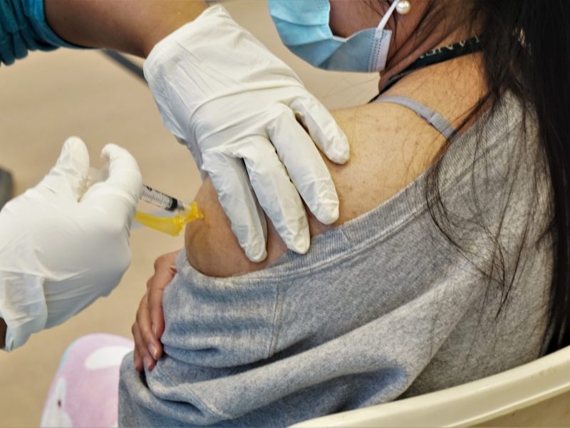 A closeup image of a young woman receiving a shot on her shoulder, with the person administering wearing latex gloves.