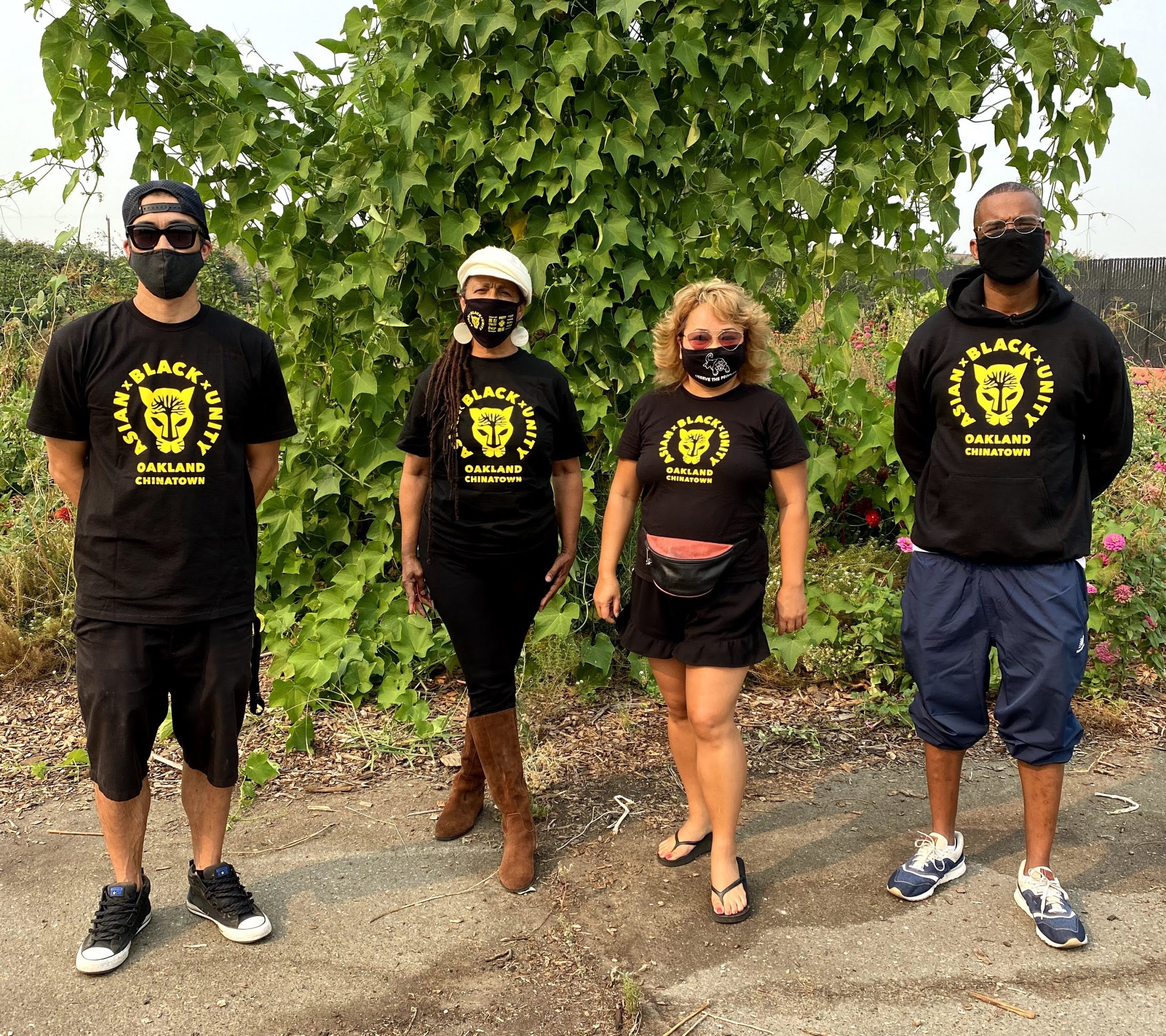 Four people stand in front of some greenery wearing Asian Black Unity t-shirts and face masks