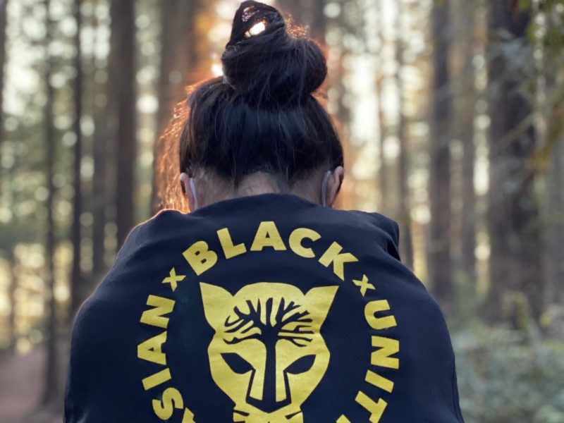 A woman in a forest wearing an Asian Black Unity black hoodie with her back facing camera