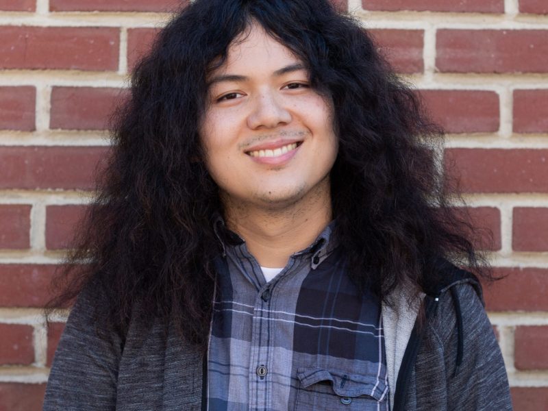 A young man with long, poofy hair stands in front of a brick wall and smiles for the camera.