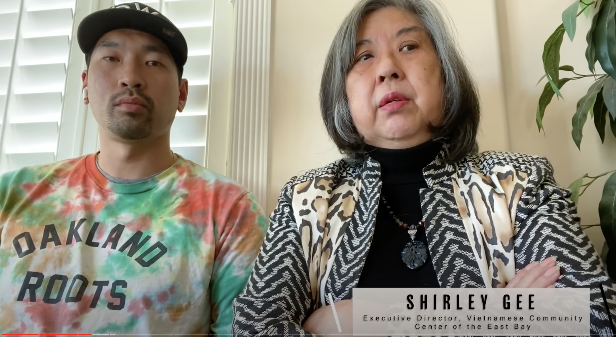 An Asian American man wearing a tie-dye shirt that says "Oakland Roots" and a woman with a bob haircut and grey hair sit together on a couch.