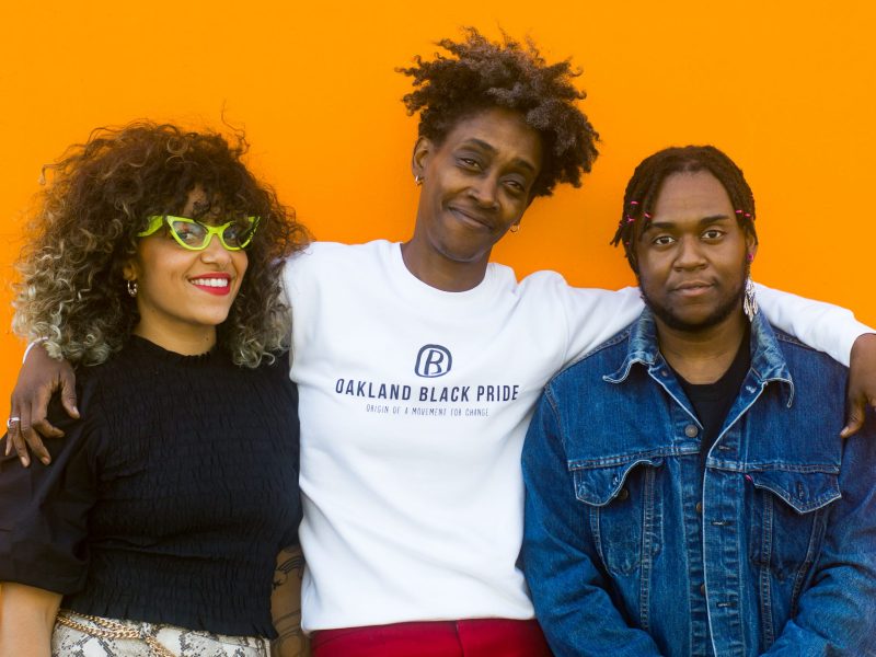 Three Black folks stand against a bright orange background with arms round each other