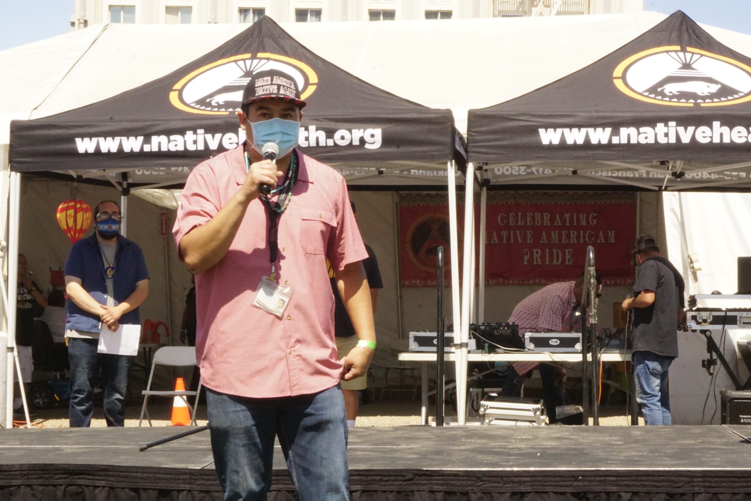 a masked Native American man speaks into a mic in front of a tent that says Native American Health Center