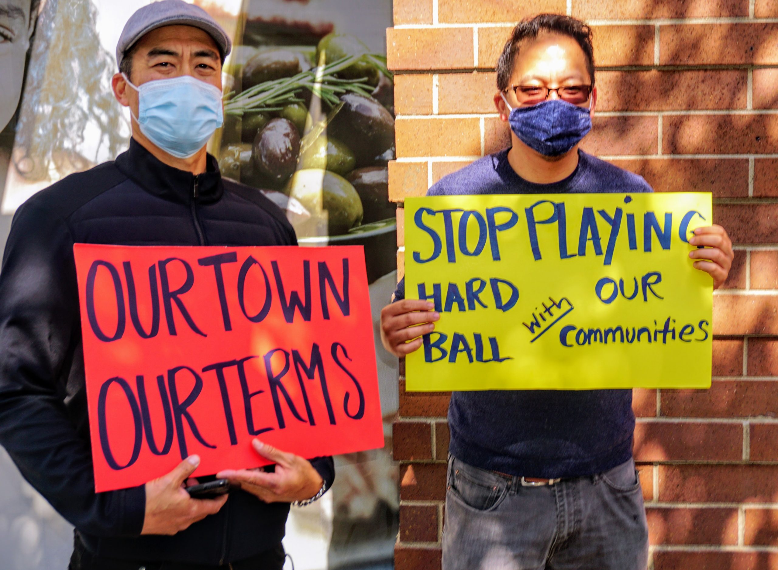 two Asian American men wearing masks hold signs next to a building that say "our town our terms."