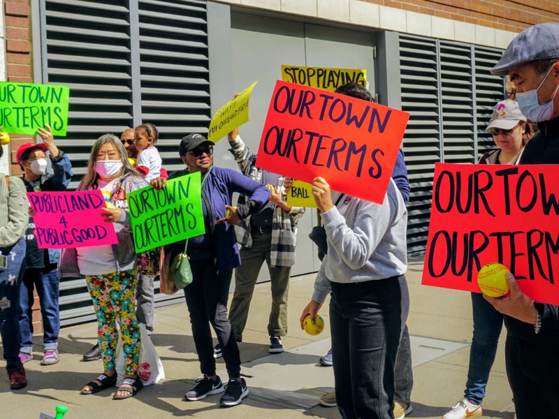 a group of people rally outside a building holding colorful signs with baseball puns asking for benefits to the community