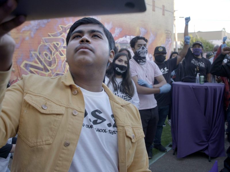 A Latinx youth holds up a screen to take a selfie with a group of people behind him smiling and posing.