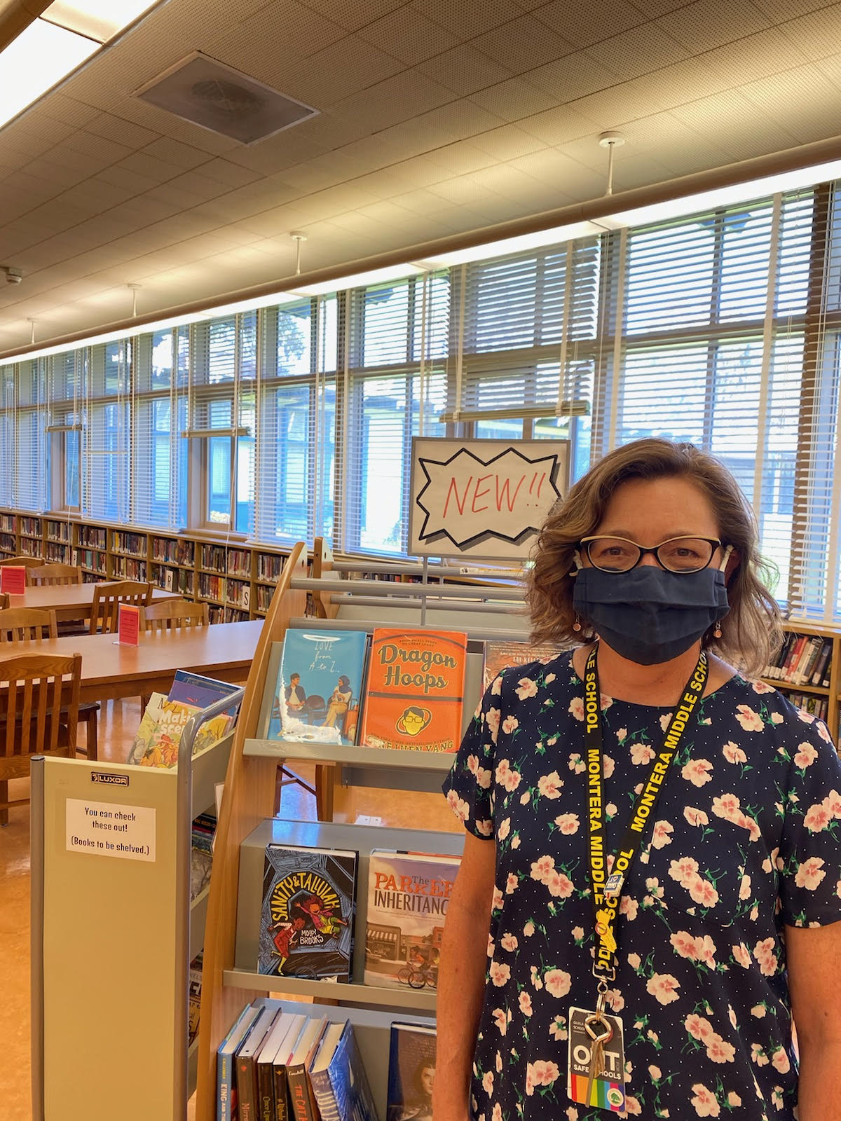 A woman wearing glasses and a mask stands inside a school library.