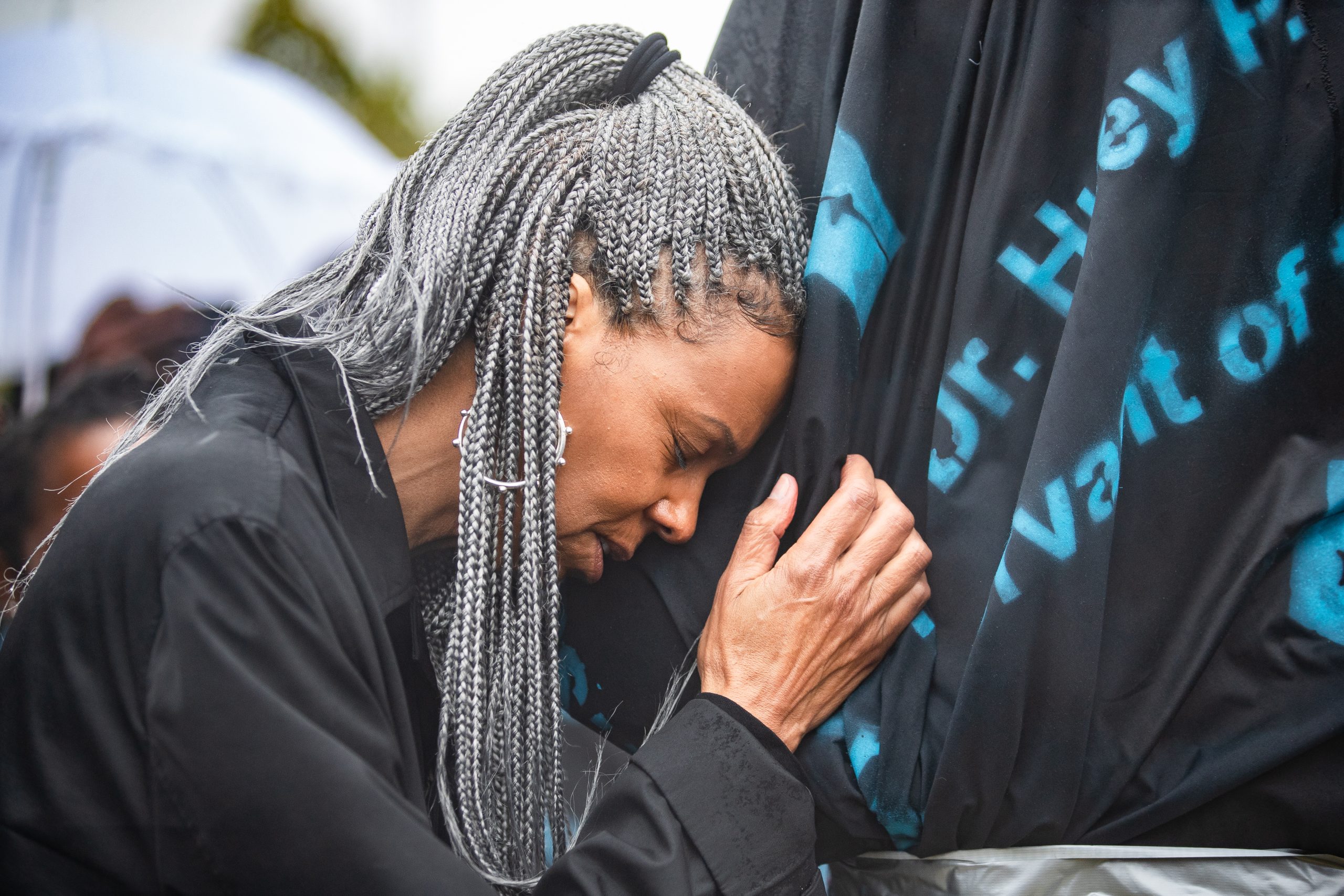 a beautiful woman with long grey braids puts her forehead on a cloth-covered statue