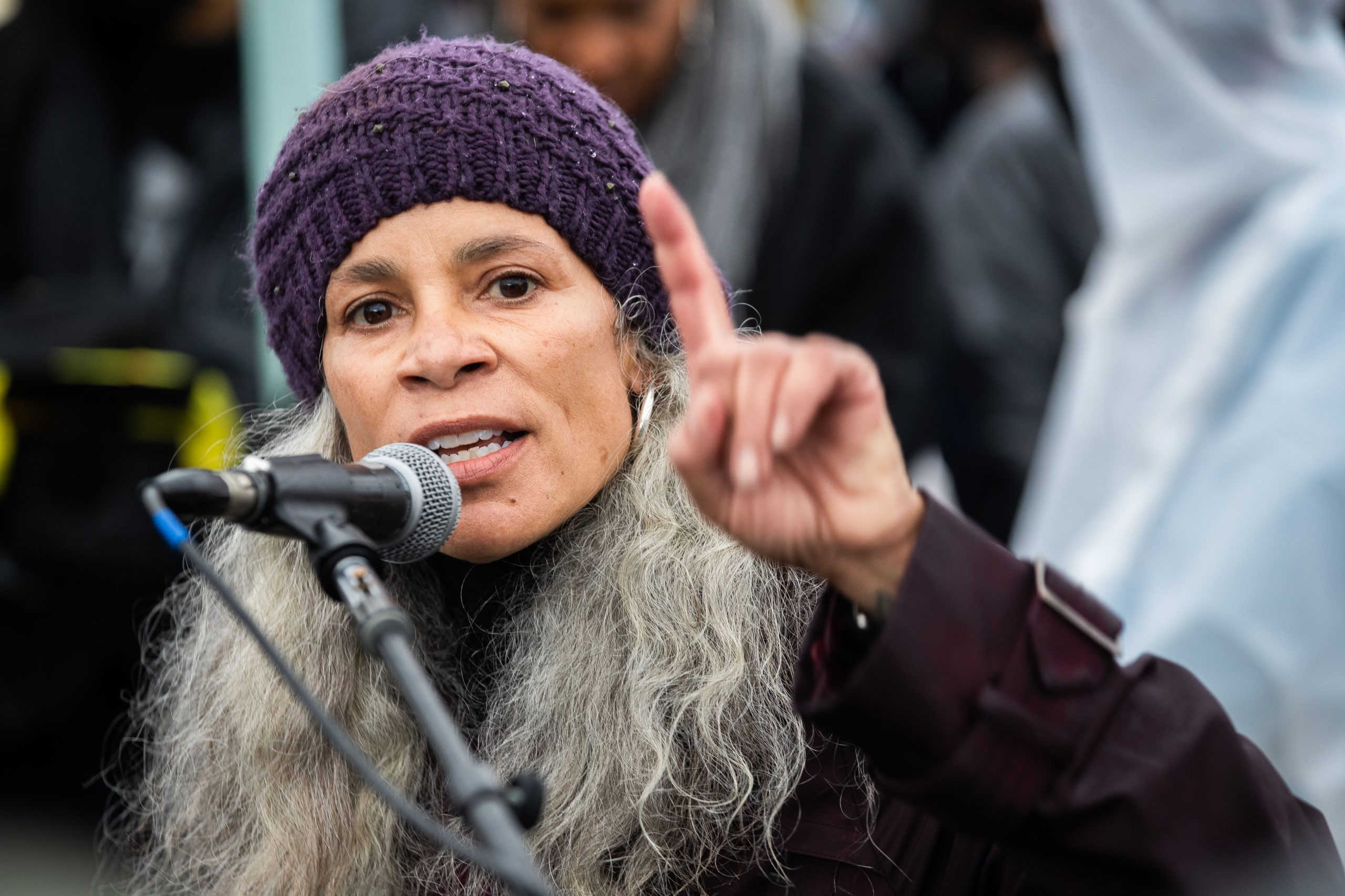 A woman wearing a crocheted purple hat and voluminous grey, curly hair speaks at the podium.